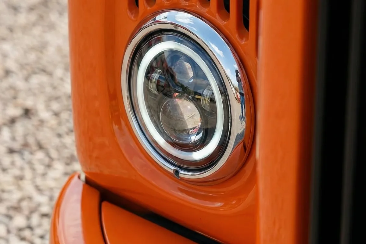 Close-up do farol dianteiro de LED circular com anel halo e moldura cromada da van customizada Scania 111S laranja.