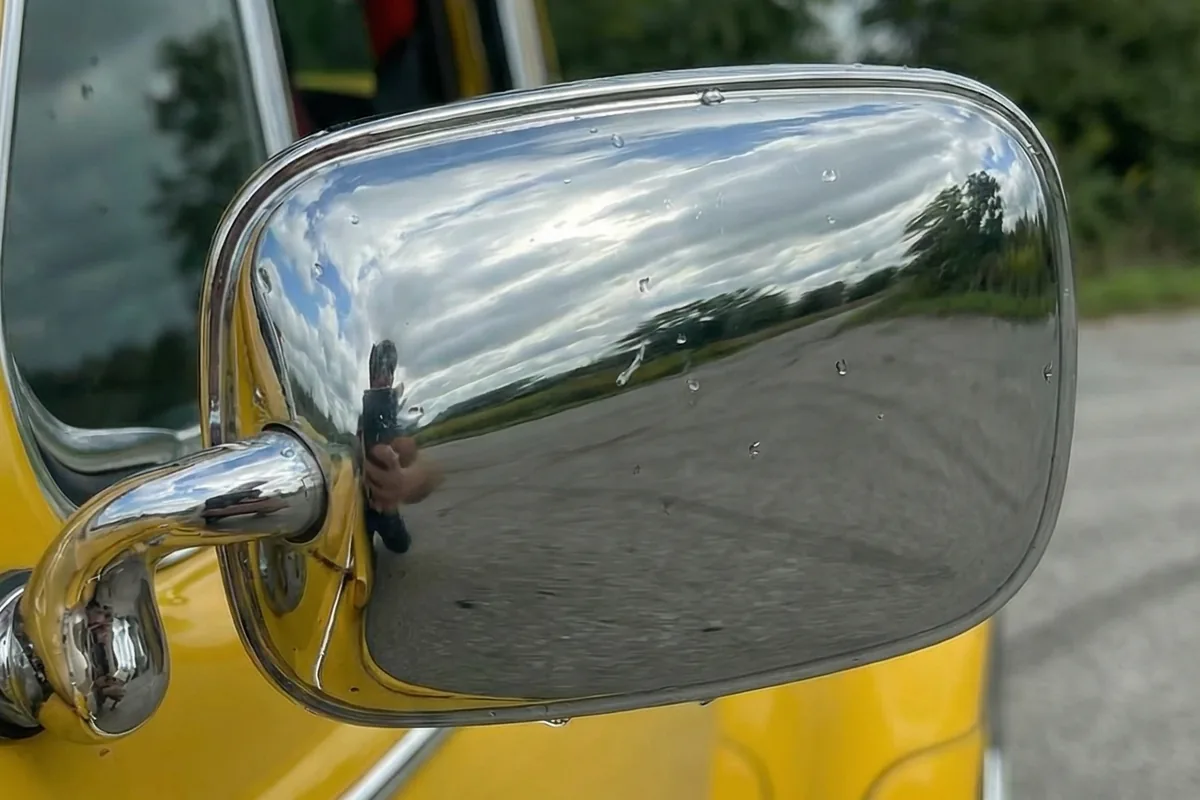 Close-up do retrovisor lateral cromado do Fusca 911 S, refletindo céu nublado, árvores e a imagem do fotógrafo.