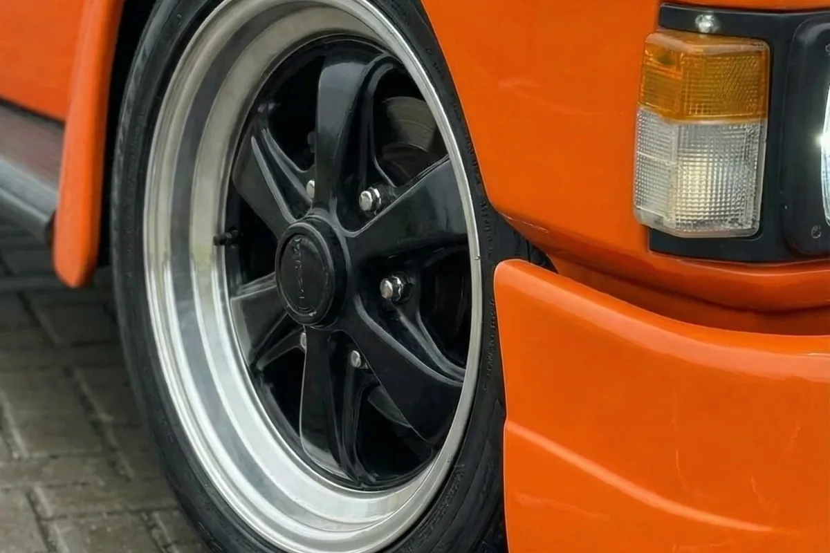Close-up da roda dianteira preta com borda polida e para-lama alargado do Fuscania 112H laranja.
