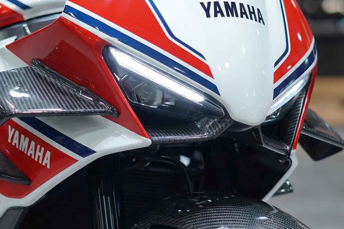 Frente da Yamaha RD350 2026 Restomod branca, vermelha e azul, faróis de LED acesos, detalhes em fibra de carbono.
