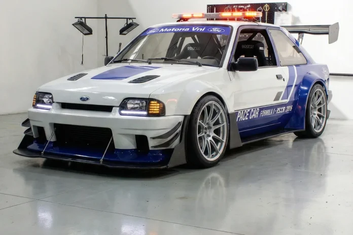 Escort-XR3-Pace-Car-Formula-1-Concept-2026-01 Ford Escort XR3 em conceito pace car, branco e azul, visto em três quartos dianteiro num galão de piso polido.