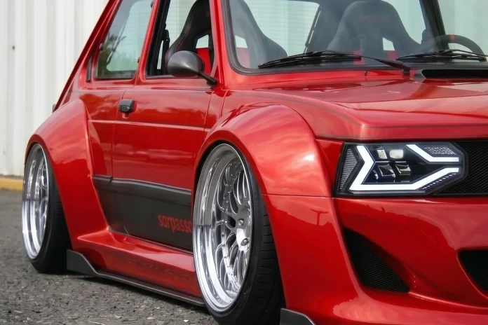 Fiat 147 vermelho em conceito widebody, visto em três quartos dianteiro junto a um galpão de chapa metálica.