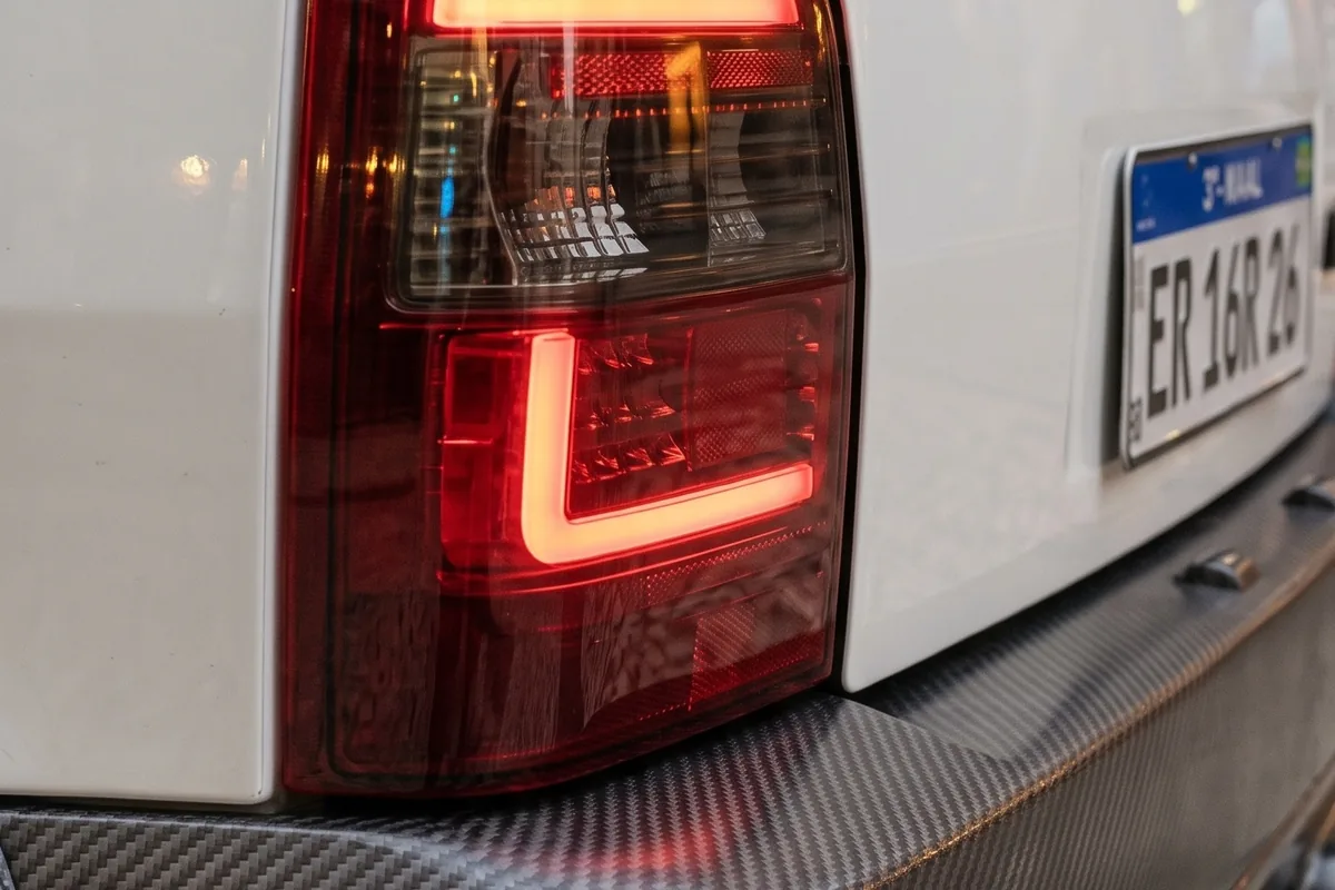 Lanterna traseira em L com LED vermelho aceso, sobre carroceria branca e difusor de fibra de carbono.