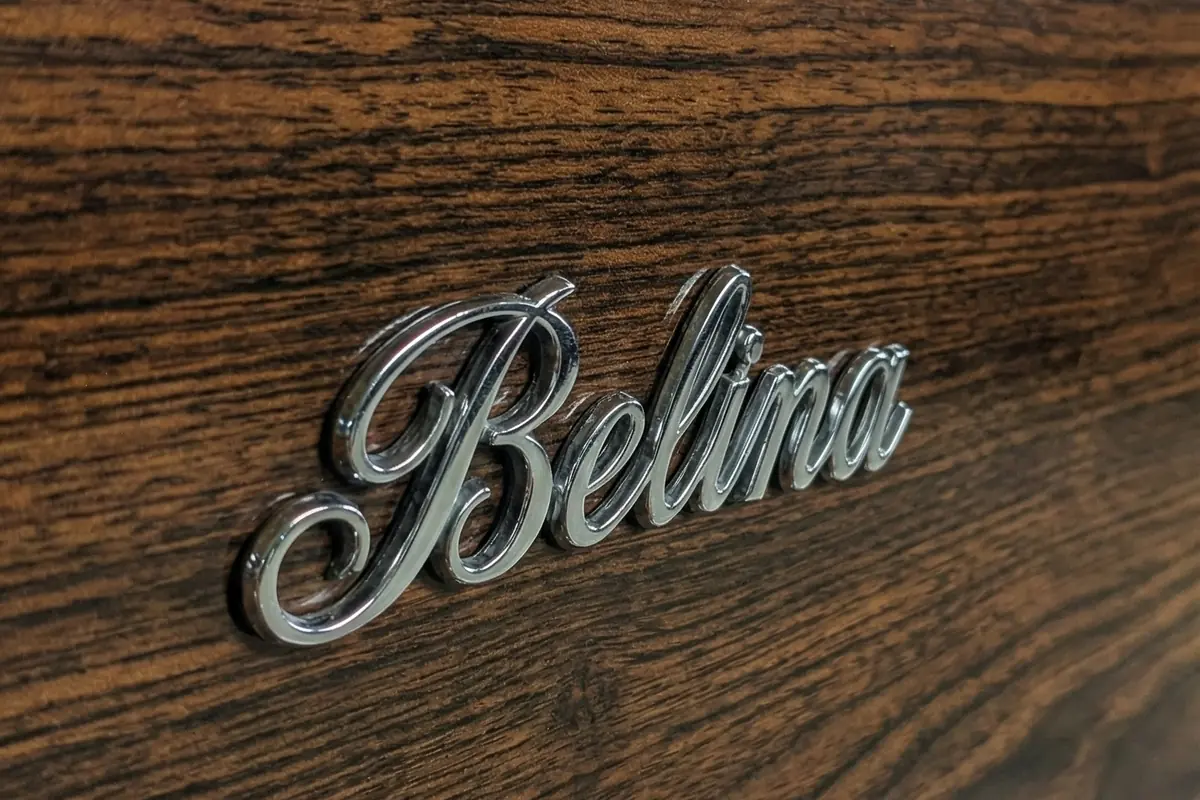 Aplique de madeira escura com friso cromado e o script Belina em metal aplicado por cima.
