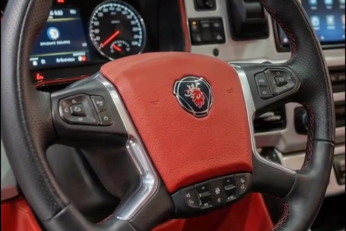 Volante vermelho e preto com emblema Scania ao centro, acompanhado por painel digital ao fundo.